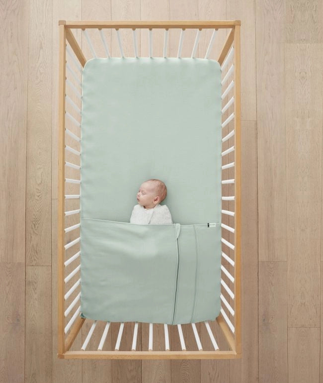 A baby sleeping blissfully on a Tuck Sheet - Sage (Cot)